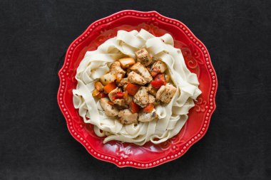 Kırmızı tabakta siyah arka planda tavuk ve sebzeli spagetti.