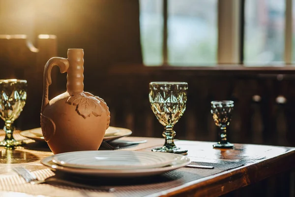 Empty reserved table in restaurant with clay vase for wine, drinking glass and plates