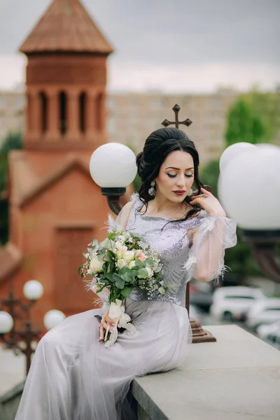 Ermeni dindar kilisesinin önünde oturan çok çekici bir nedime, gelin buketi tutuyor ve düğün töreninden sonra kameraya poz veriyor. Nedime fotoğrafı.