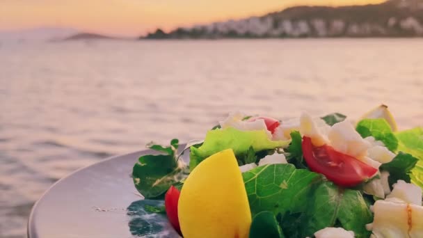 Mediterraner Salat mit weißem Käse und frischem grünen Gemüse auf dem Teller in einem Restaurant am Meer bei Sonnenuntergang — Stockvideo