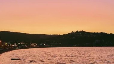 Deniz manzarası, seyahat ve doğa konsepti, deniz dalgaları ve kıyı kasabasının ışıkları gün batımında