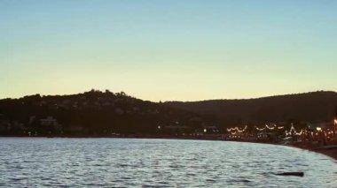 Deniz manzarası, seyahat ve doğa konsepti, deniz dalgaları ve kıyı kasabasının ışıkları gün batımında