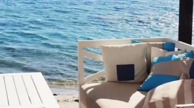 Plaj salonu ve yaz tatili konsepti. Rahat yastıkları, yastıkları ve mavi denizin yanındaki masalı beyaz kanepe.