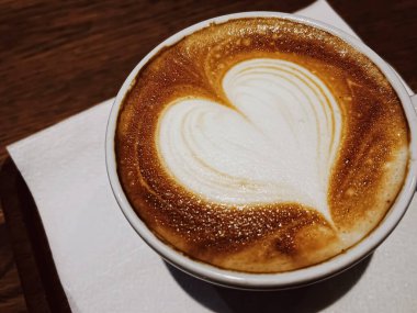 Kahve molası ve romantik ruh hali kavramı. Bir bardak karamelli kapuçino, kalp şeklinde, laktozsuz sütten yapılmış, kafeteryadaki ahşap masada servis edilen köpük sanatı.