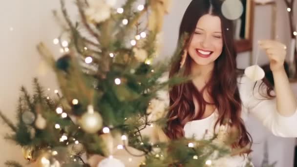 Decoración navideña y concepto de humor navideño. Feliz mujer sonriente decorando árbol de Navidad con juguetes festivos y adornos en casa, brillo brillante efecto bokeh — Vídeos de Stock