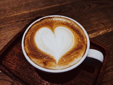 Kahve molası ve romantik ruh hali kavramı. Bir bardak karamelli kapuçino, kalp şeklinde, laktozsuz sütten yapılmış, kafeteryadaki ahşap masada servis edilen köpük sanatı..