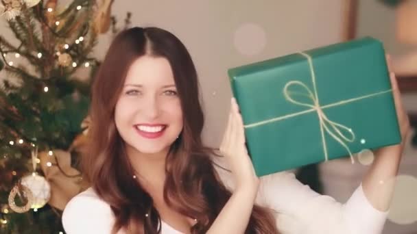 Vacaciones de Navidad y regalo para su concepto. Feliz mujer sonriente sosteniendo envuelto caja de regalo en casa, árbol de Navidad en el fondo y brillo brillante efecto bokeh — Vídeo de stock