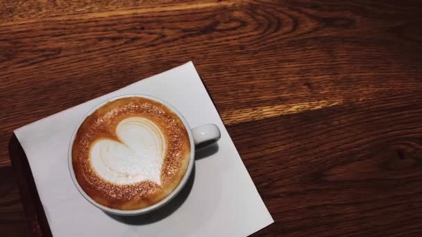 Coffee break and romantic mood concept. Cup of caramel cappuccino with heart shaped foam art made of lactose-free milk, served on wooden table in cafeteria — Stock Video