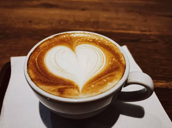 Przerwa na kawę i romantyczny nastrój. Kielich karmelowego cappuccino z piankową sztuką w kształcie serca z mleka bez laktozy, podawany na drewnianym stole w stołówce — Zdjęcie stockowe