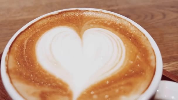 Coffee break and romantic mood concept. Cup of caramel cappuccino with heart shaped foam art made of lactose-free milk, served on wooden table in cafeteria — Stock Video