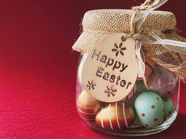 Mutlu Paskalyalar. Kırmızı arka planda cam kavanozda renkli Paskalya yumurtaları, Hıristiyan dini ve tatil kavramı