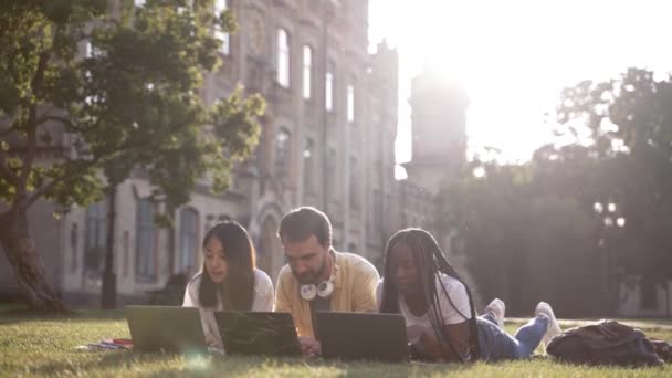 Веселі товариші з ноутбуками, що лежать на галявині кампусу — стокове відео