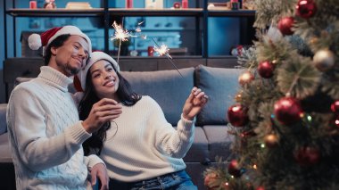 Neşeli Afrikalı Amerikalı çift Noel ağacının yanında maytap tutuyor.
