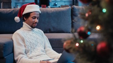 Bulanık Noel ağacının yanında dizüstü bilgisayar kullanan, Noel Baba şapkalı Afrikalı Amerikalı bir adam.