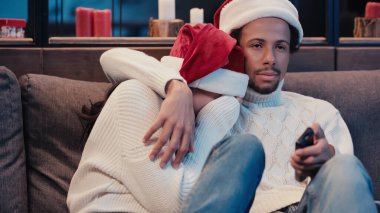 Afrikalı Amerikalı adam, korkmuş kız arkadaşıyla Noel Baba şapkası takıp film izliyor.