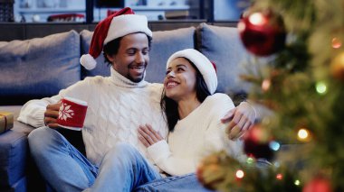 Afrika kökenli Amerikalı bir adam elinde fincanla bir kadınla Noel ağacının yanında gülümsüyor.