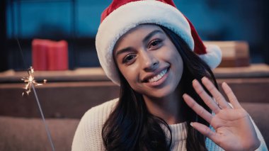 Mutlu Afro-Amerikalı kadın Noel Baba şapkalı, el sallıyor ve maytap tutuyor.