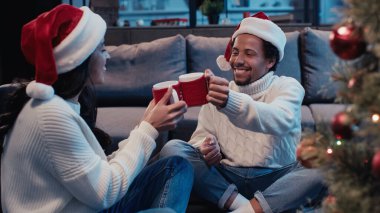 Noel Baba şapkalı mutlu Afro-Amerikan çift bulanık noel ağacının yanında kadeh tokuşturuyor. 