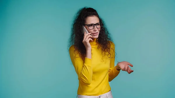 戴眼镜的年轻女人很不高兴 她用蓝色隔离的智能手机说话 — 图库照片