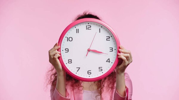 young woman obscuring face with wall clock isolated on pink