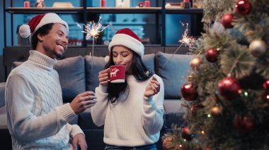 Noel Baba şapkalı ve maytaplı mutlu Afro-Amerikalı kadın. Yanında erkek ve bulanık Noel ağacı. 