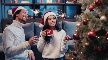 Noel Baba şapkalı neşeli Afro-Amerikalı çift ellerinde bardak ve maytap tutarken bulanık Noel ağacına bakıyorlar. 