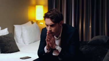 Endişeli bir işadamı otel odasında dua eden elleriyle oturuyor.