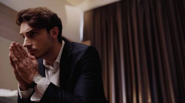 Endişeli işadamı dua eden elleriyle otel odasında oturuyor ve düşünüyor.