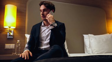 Yatakta oturan ve otel odasında akıllı telefondan konuşan iş adamı.