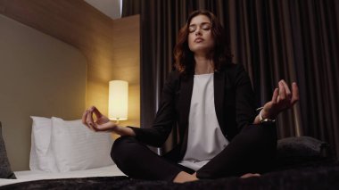 young businesswoman meditating in lotus pose in hotel room clipart