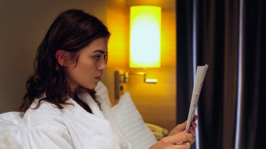 Otel odasında gazete okuyan bornozlu genç bir kadının yan görüntüsü.