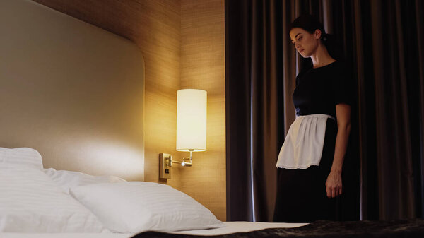 young maid in uniform and apron looking at bed in hotel room