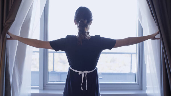 back view of young maid opening dark curtains in hotel room