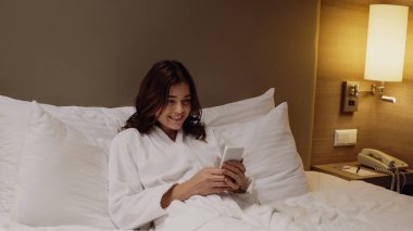 Bornoz giymiş genç, neşeli bir kadın yatakta dinlenirken akıllı telefonuyla mesajlaşıyor.