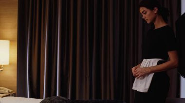 Otel odasında yatağın yanında üniformalı genç bir hizmetçi duruyor.
