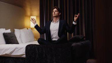 Genç işadamı, otel odasındaki yatakta nilüfer çiçeği pozuyla meditasyon yapıyor.