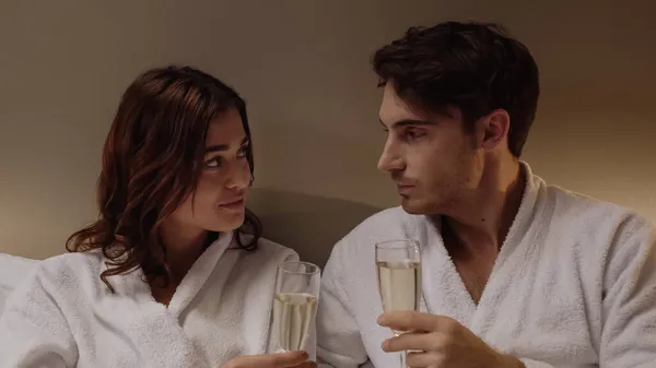 穿着浴衣的年轻夫妇一边拿着装有香槟的眼镜 一边在宾馆房间里相互望着对方 — 图库照片