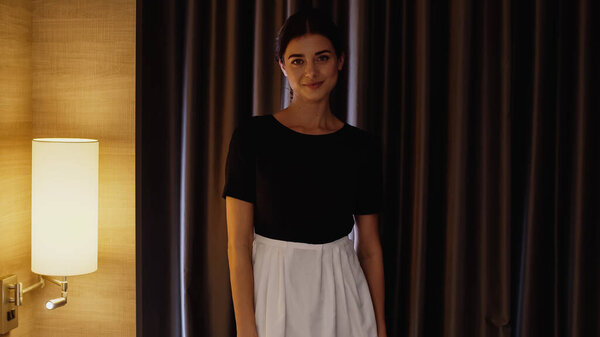 young maid in white apron smiling at camera in hotel room