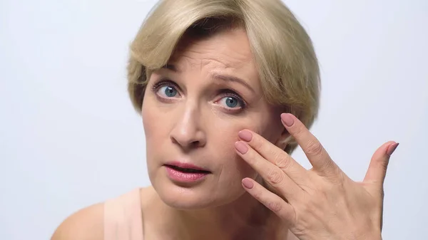 Beyazda izole edilmiş kameraya bakarken orta yaşlı bir kadın yüzüne dokunuyor.