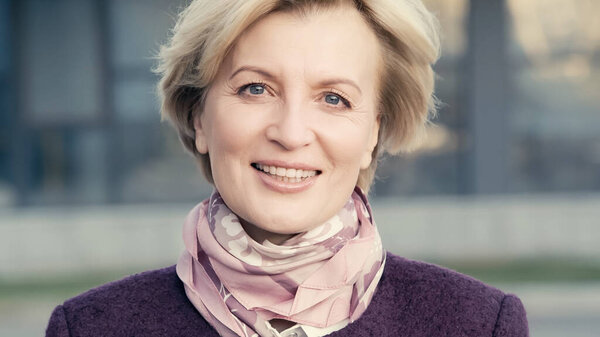 Радостная женщина средних лет в шейном шарфе смотрит в камеру на улице