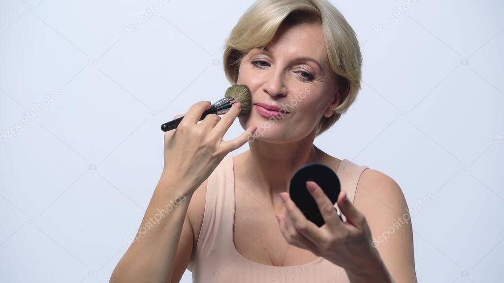 blonde mature woman applying face powder with cosmetic brush isolated on white