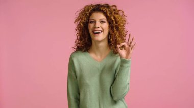excited woman with red wavy hair showing okay sign while looking at camera isolated on pink clipart