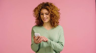 Pembe üzerine izole edilmiş cep telefonu kullanan kıvırcık saçlı neşeli bir kadın.