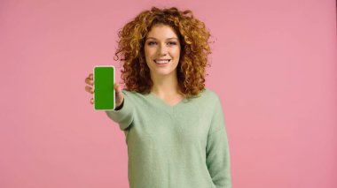Pembe ekranda izole edilmiş yeşil ekranlı akıllı telefonu gösteren neşeli kızıl saçlı kadın.
