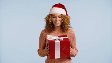 happy redhead woman in santa hat looking at new year present isolated on blue clipart