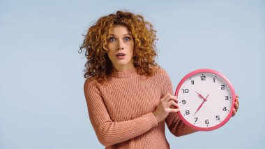 shocked redhead woman with round clock looking at camera isolated on blue clipart