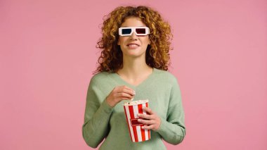 Üç boyutlu gözlüklü neşeli kadın film izliyor ve pembe üzerinde izole edilmiş patlamış mısır yiyor.