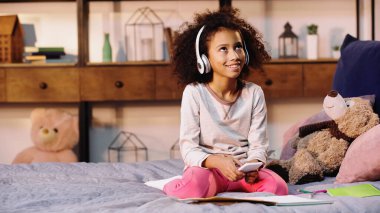 Afro-Amerikan çocuk elinde akıllı telefonuyla yatağın yanında müzik dinliyormuş.