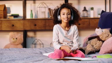 Neşeli Afro-Amerikan çocuğu, elinde defterlerle müzik dinlerken yatağın yanında akıllı telefon tutuyor.