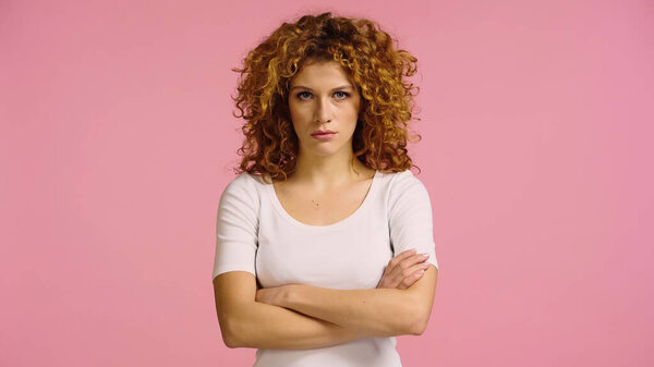 displeased redhead woman standing with crossed arms and looking at camera isolated on pink